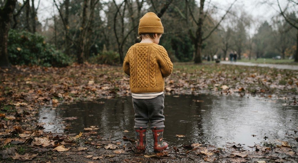 A child in layered fall clothing standing at a puddle's edge on a gray Portland day
